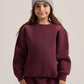 A young girl wearing a matching burgundy sweatshirt and sweatpants set with a light gray knit beanie, standing against a neutral light gray background.