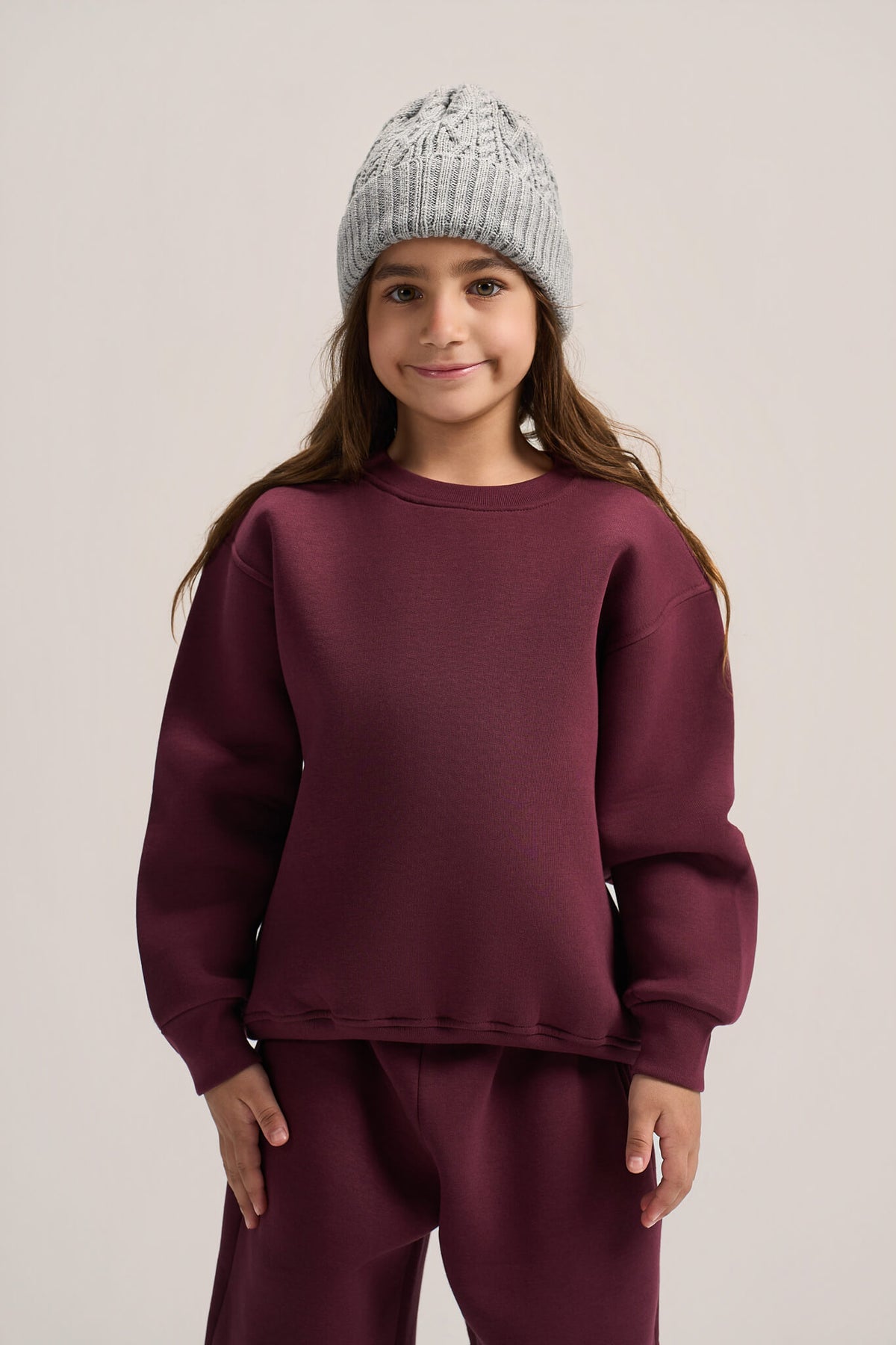 A young girl wearing a matching burgundy sweatshirt and sweatpants set with a light gray knit beanie, standing against a neutral light gray background.