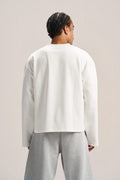 a man showing his back profile wearing a white long sleeves boxy t-shirt on an ash grey sweatpants with a solid background  