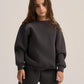 A young girl with long brown hair wearing a dark charcoal gray sweatshirt and matching sweatpants, standing against a light beige studio background.