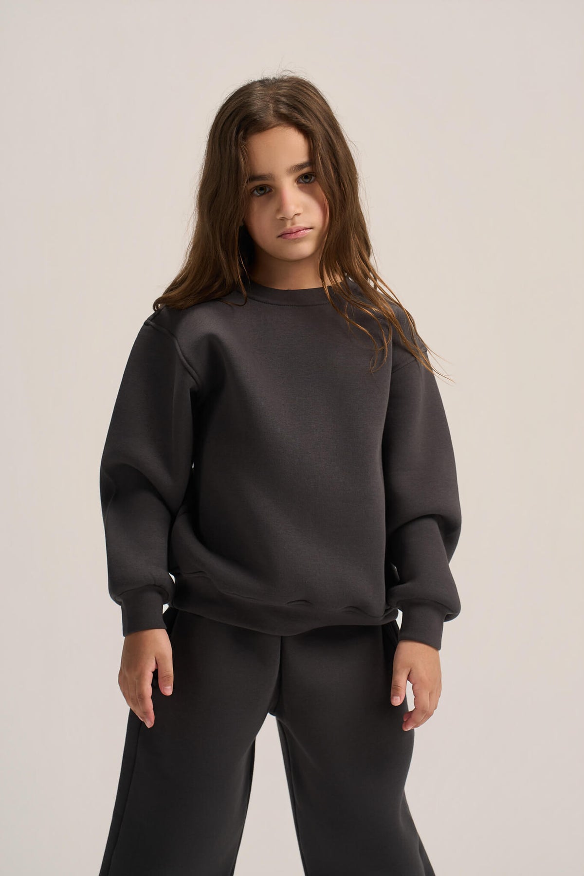 A young girl with long brown hair wearing a dark charcoal gray sweatshirt and matching sweatpants, standing against a light beige studio background.