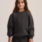 A young girl with long brown hair wearing a dark charcoal gray crewneck sweatshirt and matching dark pants, standing against a light gray studio background.