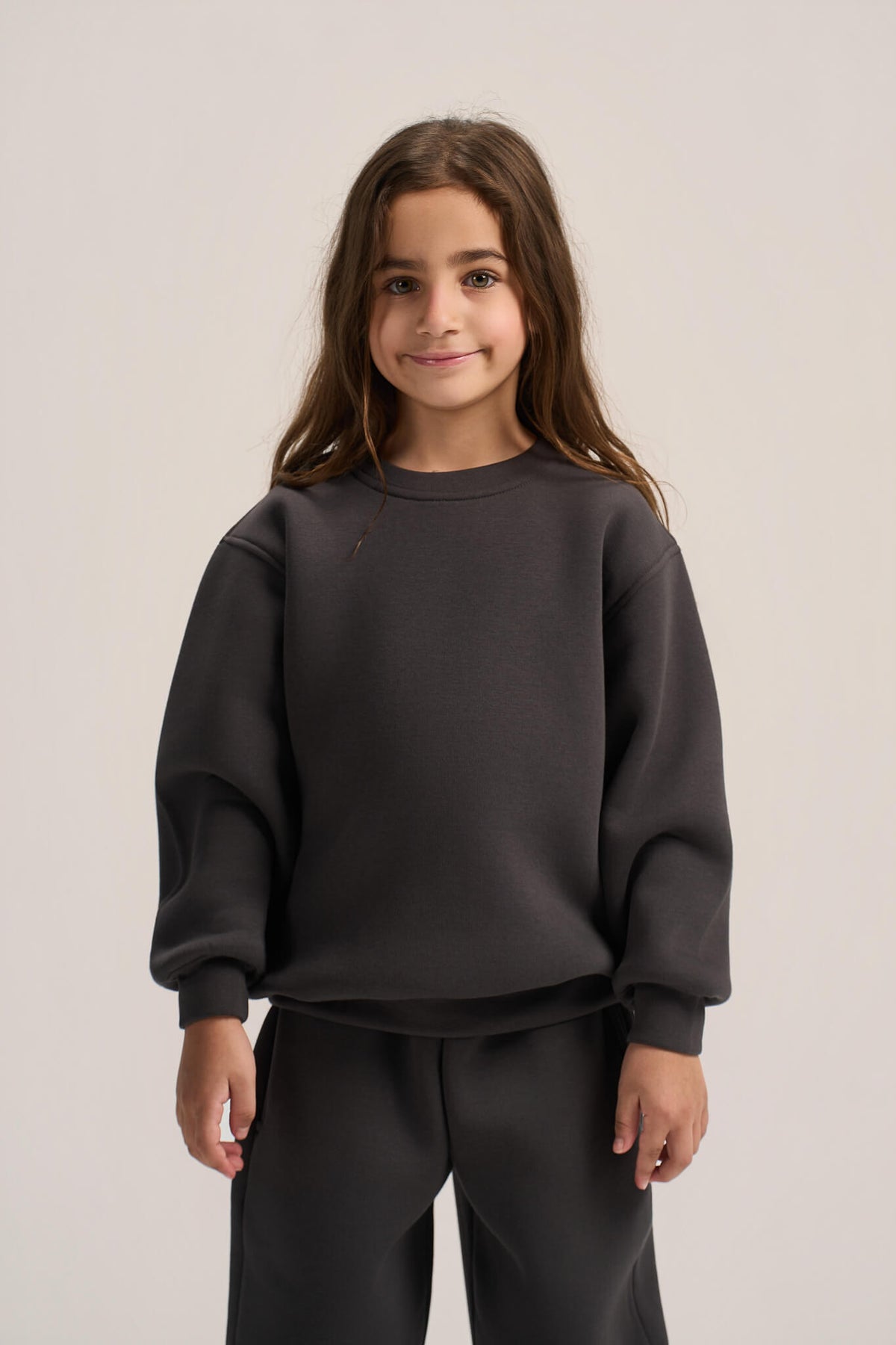 A young girl with long brown hair wearing a dark charcoal gray crewneck sweatshirt and matching dark pants, standing against a light gray studio background.