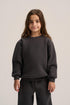 A young girl with long brown hair wearing a dark charcoal gray crewneck sweatshirt and matching dark pants, standing against a light gray studio background.