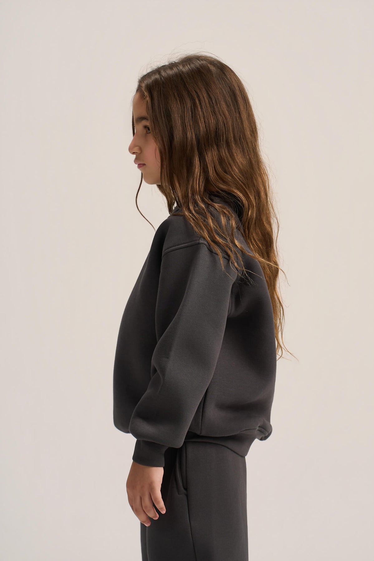 A side-profile view of a young girl with long brown hair wearing a dark charcoal gray sweatshirt and matching sweatpants against a plain light beige studio background.