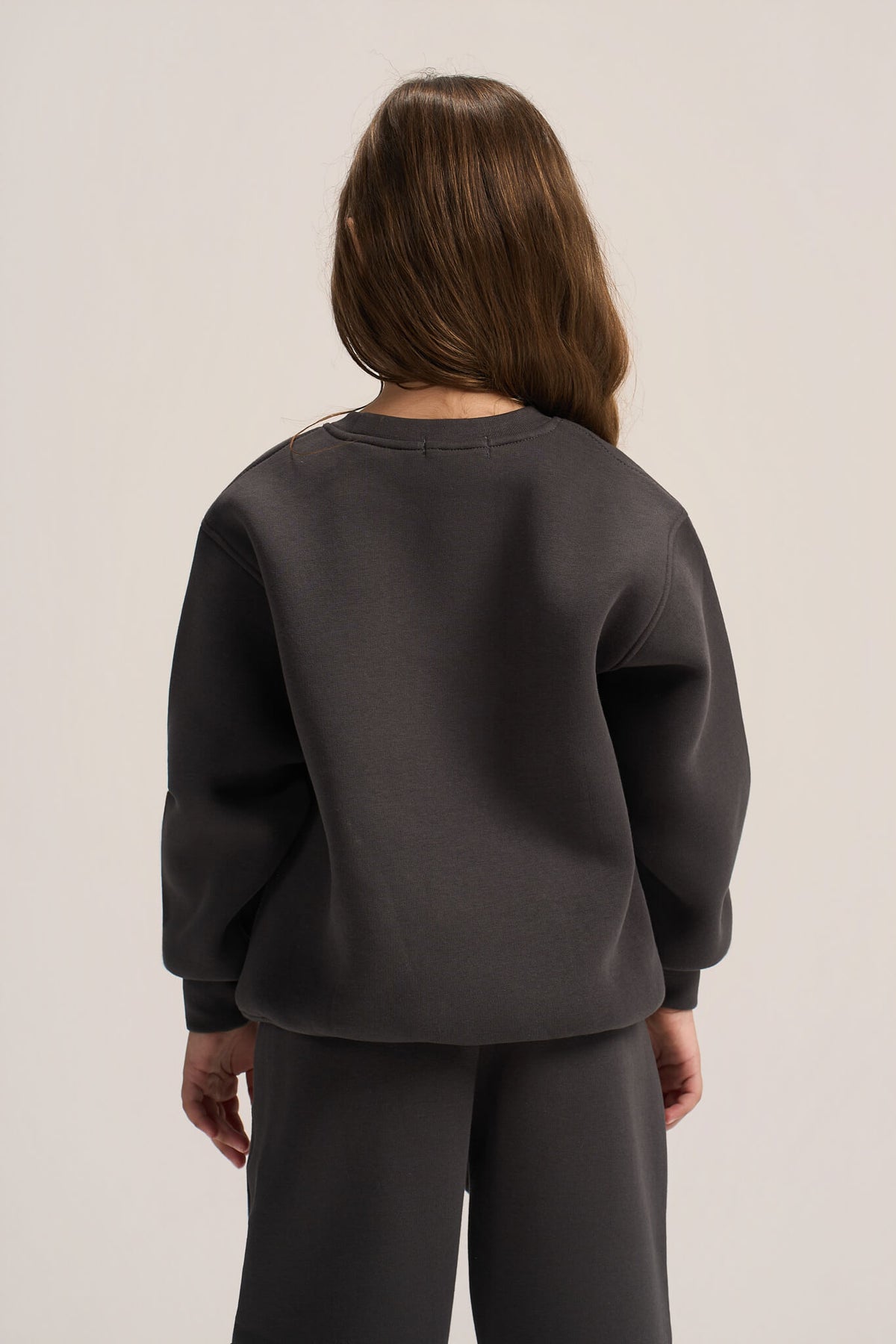 A model shown from the back wearing a dark charcoal gray crewneck sweatshirt and matching dark gray sweatpants against a light gray studio background.