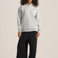 A woman modeling a light gray heathered pullover hoodie and black wide-leg pants against a solid light beige studio background.