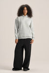 A woman modeling a light gray heathered pullover hoodie and black wide-leg pants against a solid light beige studio background.