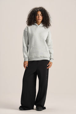 A woman modeling a light gray heathered pullover hoodie and black wide-leg pants against a solid light beige studio background.