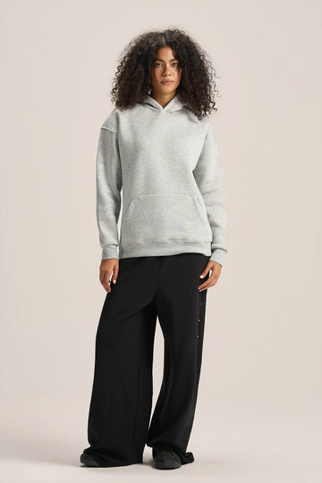 A woman modeling a light gray heathered pullover hoodie and black wide-leg pants against a solid light beige studio background.