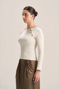 A woman modeling a fitted long-sleeve cream top with decorative gold-toned buttons along the neckline, paired with brown draped trousers, against a plain light beige studio background.