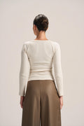 A woman shown from the back wearing a fitted cream ribbed long-sleeve top and brown wide-leg pants against a plain light beige studio background.