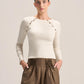 A woman modeling a fitted cream long-sleeve top with decorative gold-tone chain and bead detailing across the shoulders, paired with pleated brown wide-leg pants, against a plain light beige studio background.