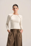 A woman modeling a fitted cream long-sleeve top with decorative gold-tone chain and bead detailing across the shoulders, paired with pleated brown wide-leg pants, against a plain light beige studio background.