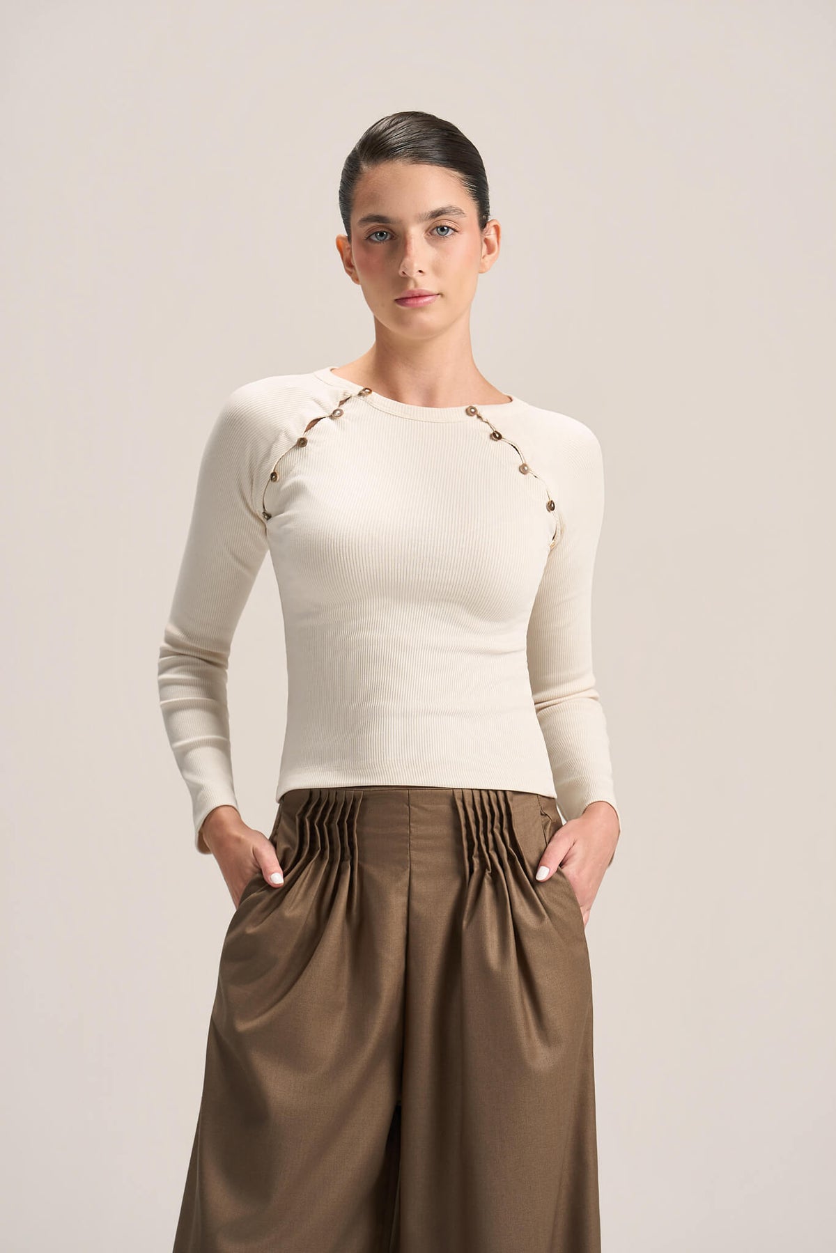 A woman modeling a fitted cream long-sleeve top with decorative gold-tone chain and bead detailing across the shoulders, paired with pleated brown wide-leg pants, against a plain light beige studio background.
