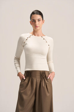 A woman modeling a fitted cream long-sleeve top with decorative gold-tone chain and bead detailing across the shoulders, paired with pleated brown wide-leg pants, against a plain light beige studio background.