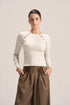 A woman modeling a fitted cream long-sleeve top with decorative gold-tone chain and bead detailing across the shoulders, paired with pleated brown wide-leg pants, against a plain light beige studio background.