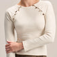 A woman wearing a cream ribbed long-sleeve top with decorative metallic buttons along the shoulders, paired with brown pleated bottoms, against a light beige studio background.