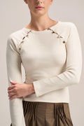 A woman wearing a cream ribbed long-sleeve top with decorative metallic buttons along the shoulders, paired with brown pleated bottoms, against a light beige studio background.
