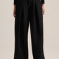 A model from the back wearing black wide-leg pleated trousers with a drawstring waist, styled with a black long-sleeve top and brown leather shoes against a light beige studio background.