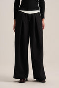 A model from the back wearing black wide-leg pleated trousers with a drawstring waist, styled with a black long-sleeve top and brown leather shoes against a light beige studio background.