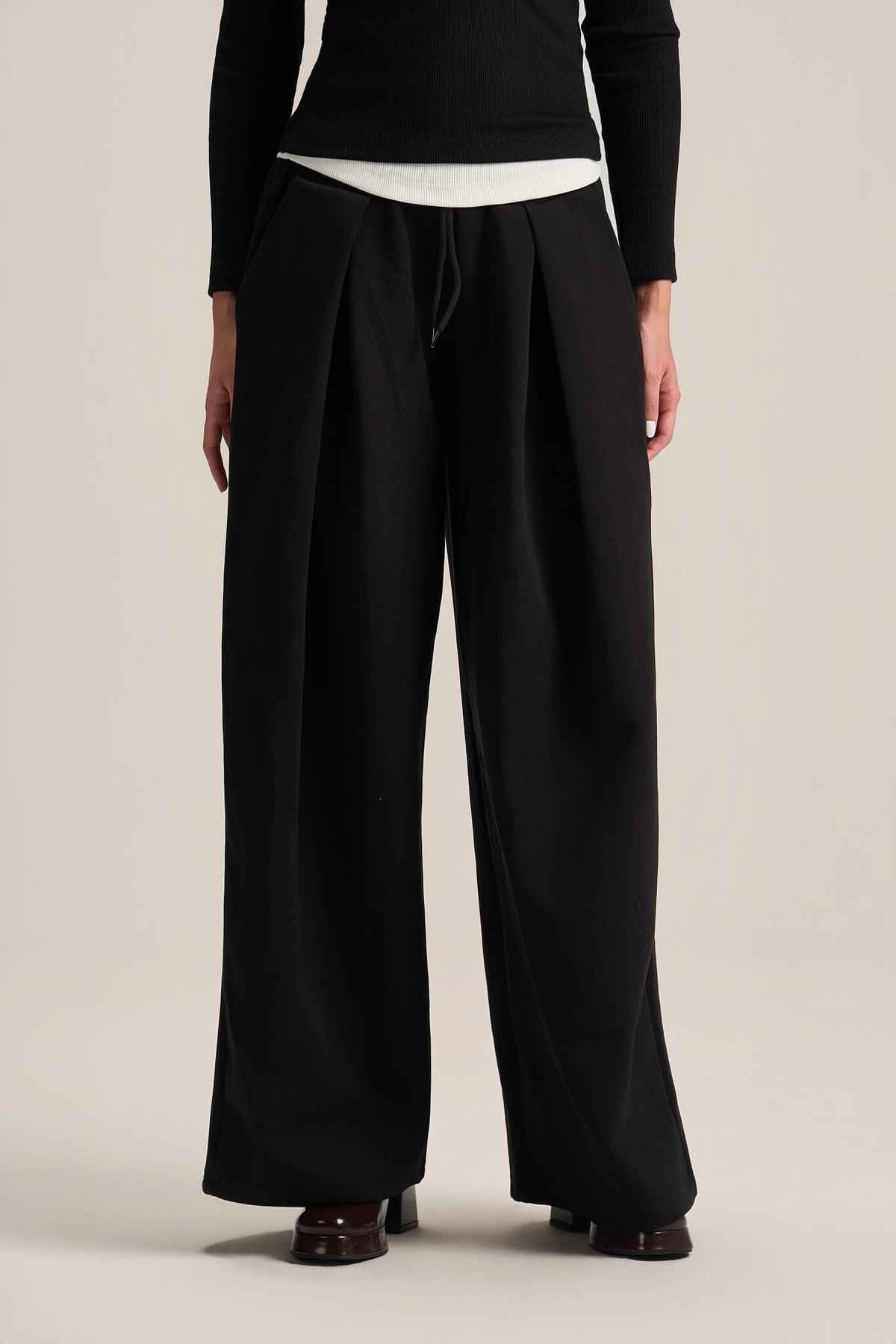 A model from the back wearing black wide-leg pleated trousers with a drawstring waist, styled with a black long-sleeve top and brown leather shoes against a light beige studio background.