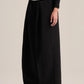 A model wearing black wide-leg trousers with a drawstring waist, styled with a black long-sleeve top and dark brown leather shoes, against a plain beige studio background.