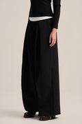 A model wearing black wide-leg trousers with a drawstring waist, styled with a black long-sleeve top and dark brown leather shoes, against a plain beige studio background.