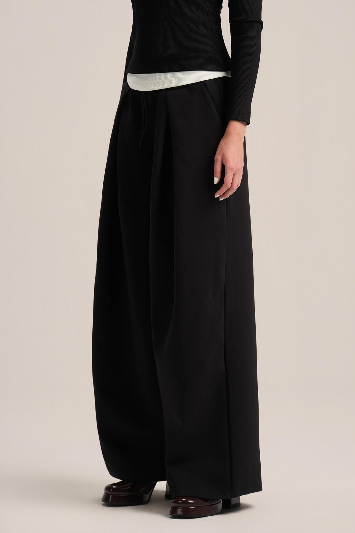 A model wearing black wide-leg trousers with a drawstring waist, styled with a black long-sleeve top and dark brown leather shoes, against a plain beige studio background.