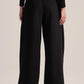 A model from the back wearing black wide-leg trousers with a contrasting white waistband, paired with a black long-sleeve top and black shoes, against a light beige studio background.