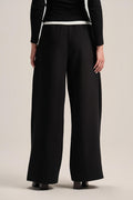 A model from the back wearing black wide-leg trousers with a contrasting white waistband, paired with a black long-sleeve top and black shoes, against a light beige studio background.
