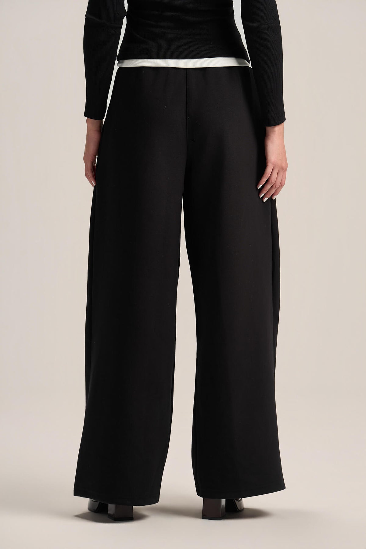 A model from the back wearing black wide-leg trousers with a contrasting white waistband, paired with a black long-sleeve top and black shoes, against a light beige studio background.