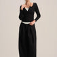 A model stands against a light beige studio background wearing a black long-sleeve knit top with a scoop neckline and button detail, paired with black wide-leg trousers.