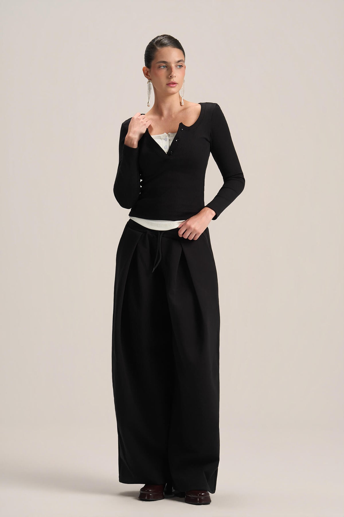 A model stands against a light beige studio background wearing a black long-sleeve knit top with a scoop neckline and button detail, paired with black wide-leg trousers.