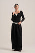 A model stands against a light beige studio background wearing a black long-sleeve knit top with a scoop neckline and button detail, paired with black wide-leg trousers.