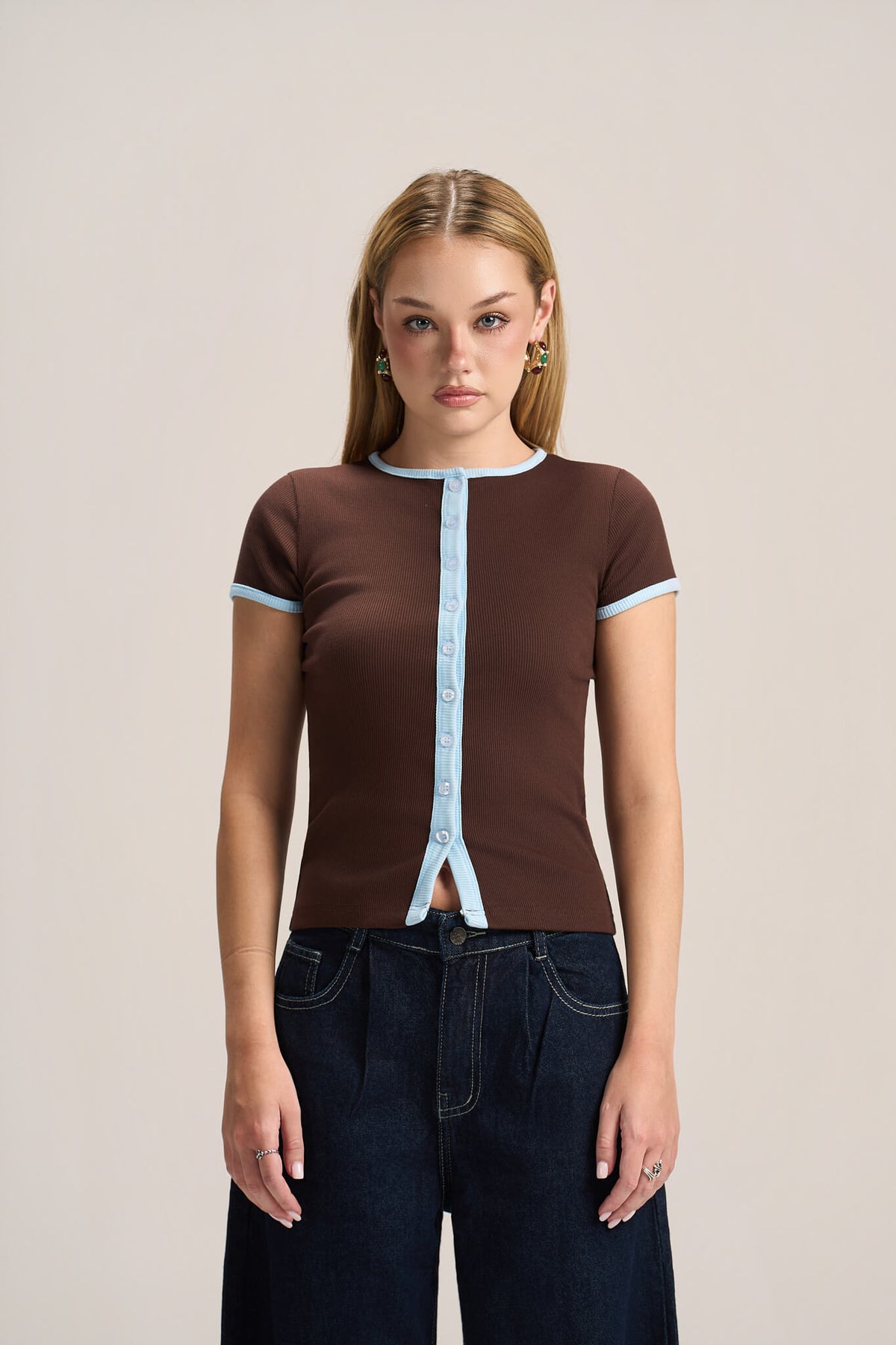A woman modeling a brown short-sleeve ribbed knit top with light blue trim details, paired with dark blue jeans, against a solid light beige background.