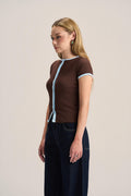 A woman modeling a brown short-sleeve knit top with light blue trim along the collar, front opening, and sleeve cuffs, paired with dark wide-leg jeans against a plain light beige studio background.
