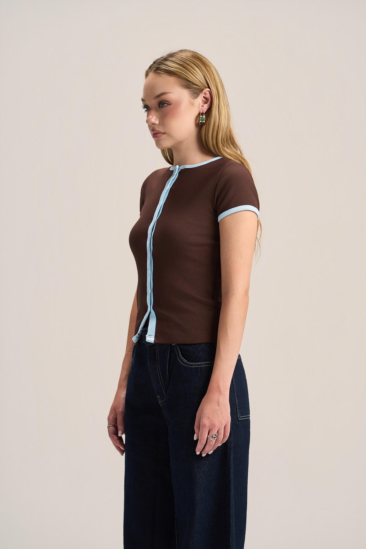 A woman modeling a brown short-sleeve knit top with light blue trim along the collar, front opening, and sleeve cuffs, paired with dark wide-leg jeans against a plain light beige studio background.
