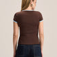 A woman viewed from the back wearing a fitted brown short-sleeve top with light blue trim on the collar and sleeve cuffs, paired with dark blue jeans, against a plain light beige studio background.