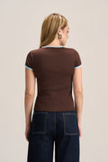 A woman viewed from the back wearing a fitted brown short-sleeve top with light blue trim on the collar and sleeve cuffs, paired with dark blue jeans, against a plain light beige studio background.