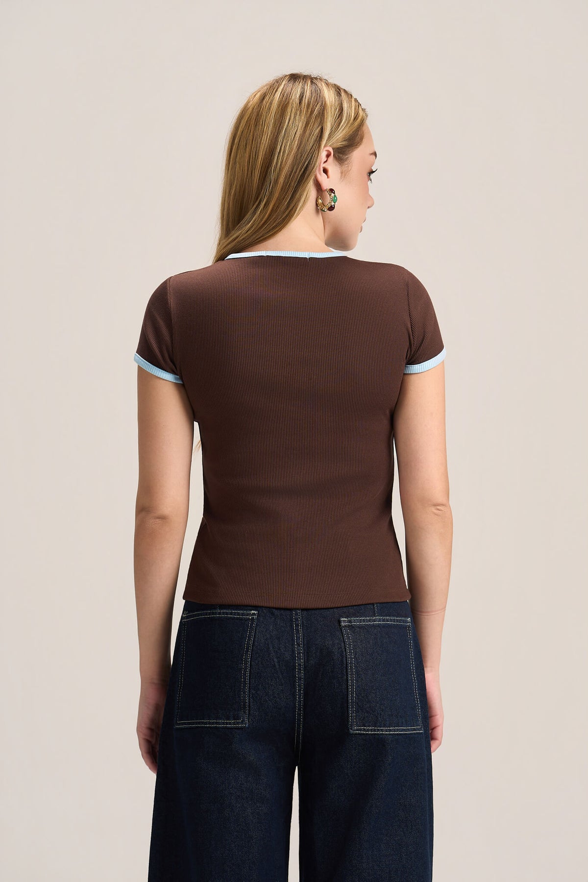 A woman viewed from the back wearing a fitted brown short-sleeve top with light blue trim on the collar and sleeve cuffs, paired with dark blue jeans, against a plain light beige studio background.