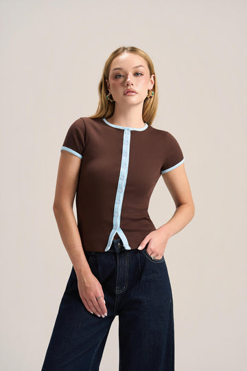 A woman modeling a brown short-sleeve ribbed top with light blue trim details, paired with dark blue jeans, against a plain light beige studio background.
