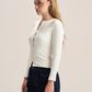 A woman with blonde hair is shown in profile wearing a fitted, long-sleeve cream-colored knit top with a split neckline and dark blue jeans, set against a plain light beige studio background.