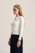 A woman with blonde hair is shown in profile wearing a fitted, long-sleeve cream-colored knit top with a split neckline and dark blue jeans, set against a plain light beige studio background.