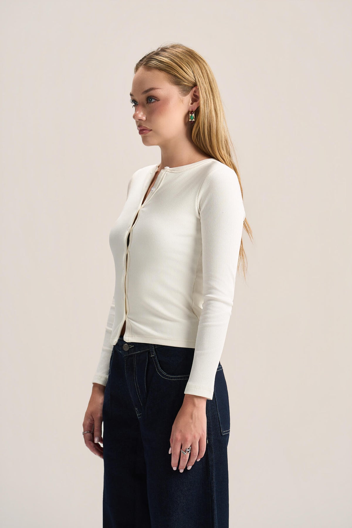 A woman with blonde hair is shown in profile wearing a fitted, long-sleeve cream-colored knit top with a split neckline and dark blue jeans, set against a plain light beige studio background.