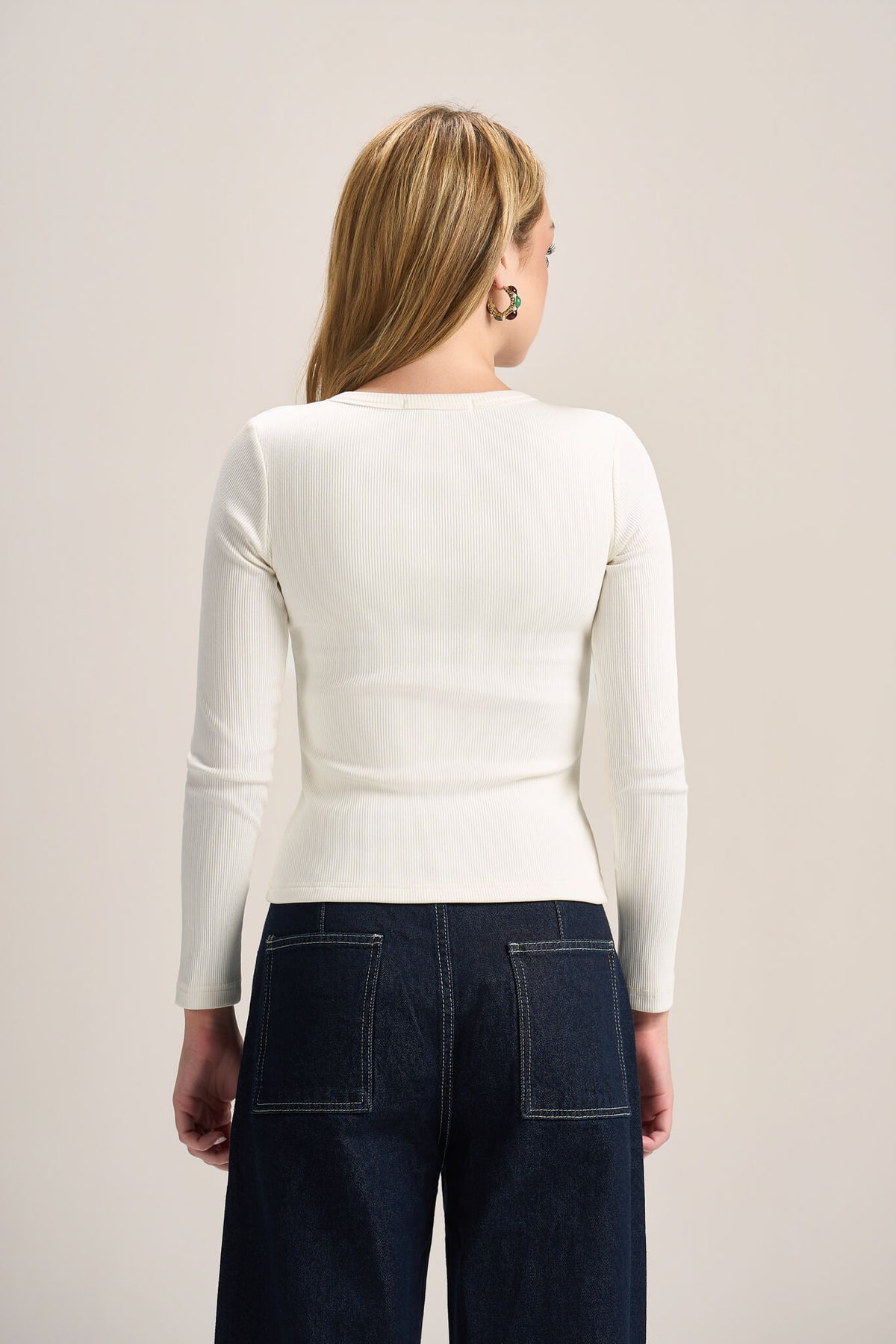 A woman shown from the back wearing a fitted off-white long-sleeve top and dark blue jeans against a plain light beige studio background.