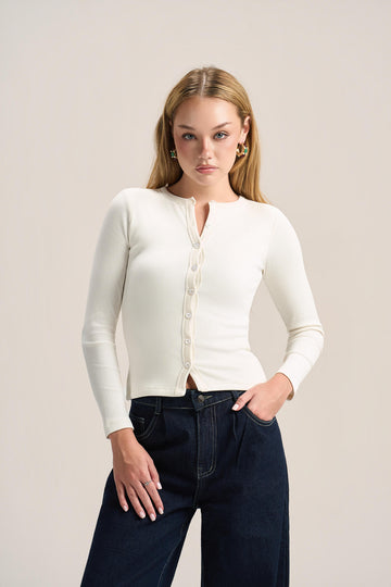 A woman modeling a fitted long-sleeve cream knit cardigan with a split neckline and front buttons, paired with dark blue jeans, against a plain light beige studio background.
