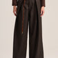 A model is shown from the waist down wearing dark brown wide-leg pleated trousers with a matching brown belt featuring a gold buckle and a braided cord detail.