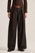 A model is shown from the waist down wearing dark brown wide-leg pleated trousers with a matching brown belt featuring a gold buckle and a braided cord detail.