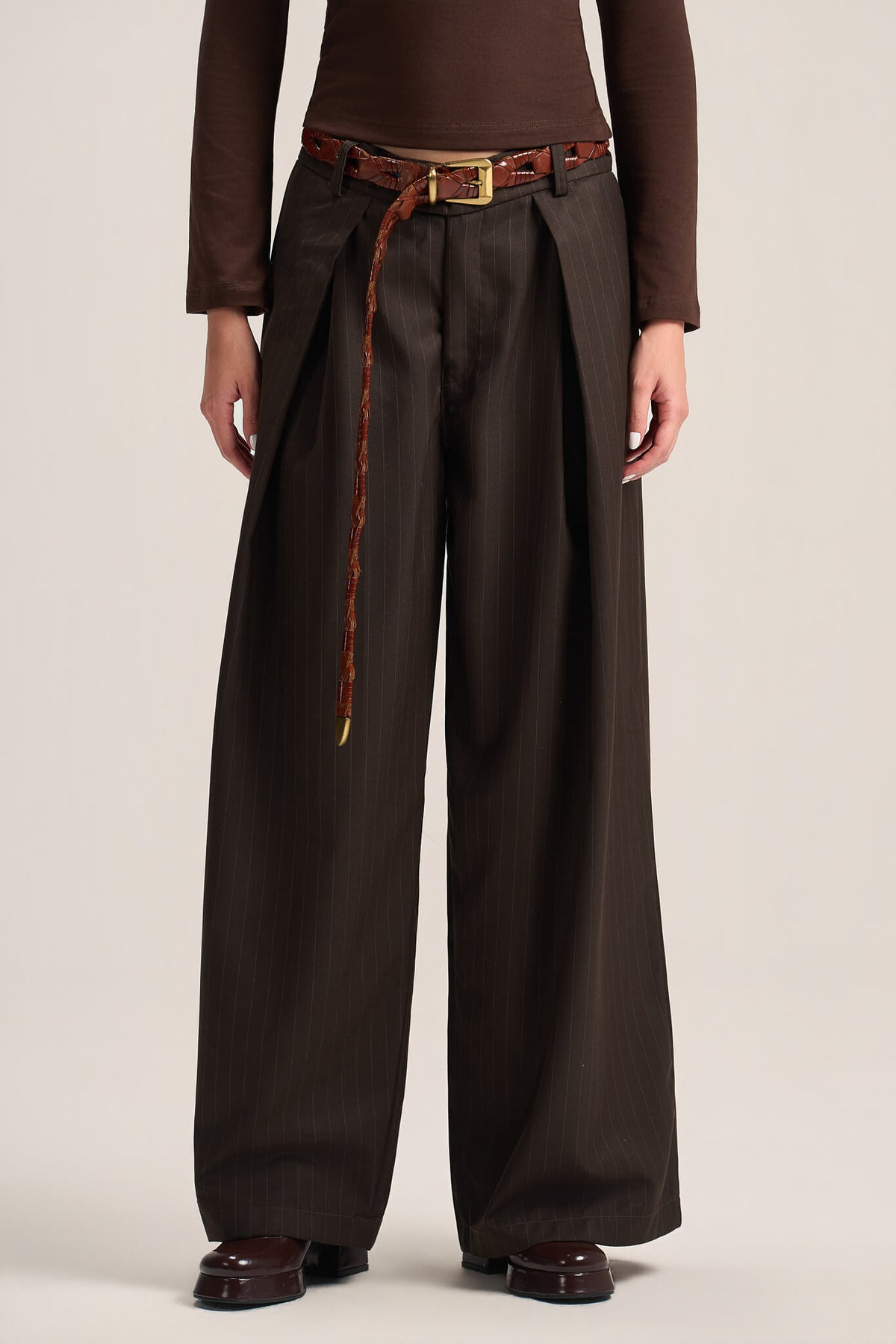 A model is shown from the waist down wearing dark brown wide-leg pleated trousers with a matching brown belt featuring a gold buckle and a braided cord detail.